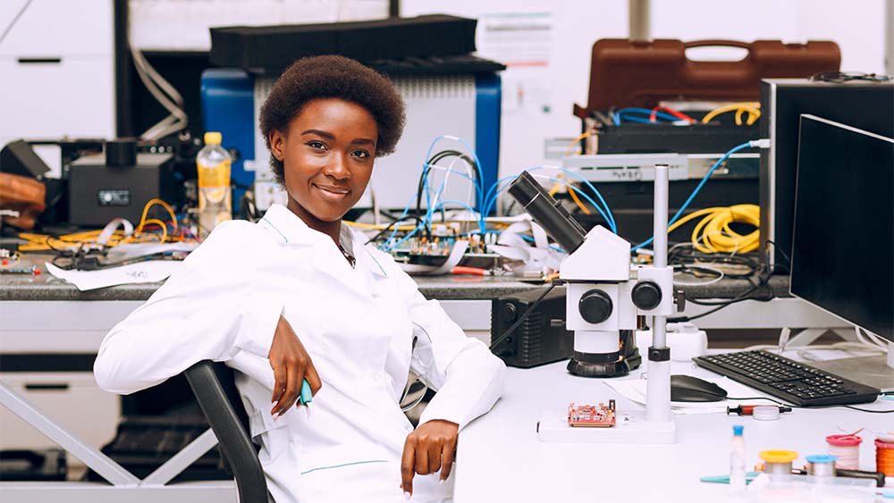 A person wearing a white coat beams at the camera. A computer and a microscope are in front of him. Numerous electronic gadgets and cables of various colors can be seen in the backdrop. 