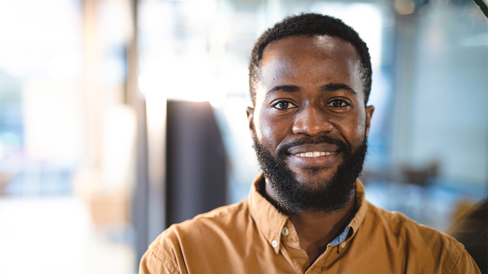 Close-up of a man looking into the camera, smiling and wearing an orange shirt in a bright setting. 