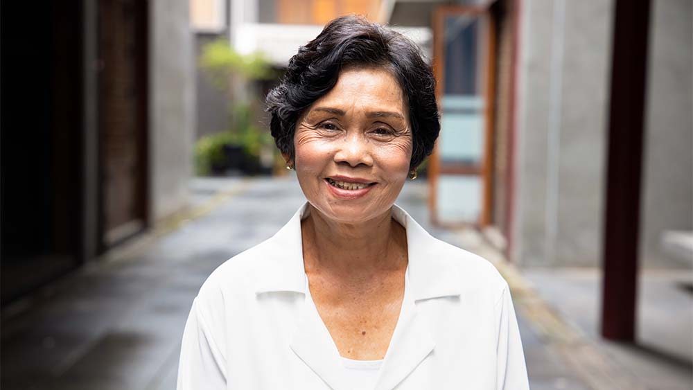 An elderly woman with a warm smile, standing in a charming alleyway. 